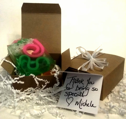 Two brown cardboard boxes, one opened with a bar of soap inside. Soap is smooth glycerin with white, pink and green swirls on a clear background. Second box is closed with a white ribbon. Small thank you card with lovely handwriting. 