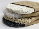 Multiple soap lifts in a stack. Each soap lift is made of compressed material  in an oval shape with a brown tag encircling the item. White, clear, tan and black color options.