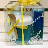 Side of gift pack, clear plastic box with yellow bow, travel sized items include blue lotion, blue body polish and full-sized blue soap bar.
